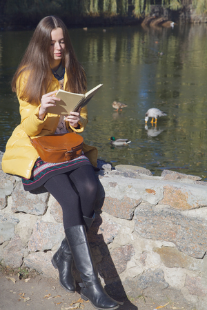 Beautiful thoughtful girl-student sits on parapet near the city pond in sunny autumn dayの写真素材