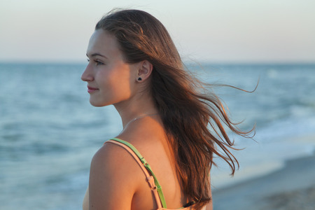 Young long-haired teen girl with brown hair standing in profile on the beach and watching to sea on a summer eveningの写真素材