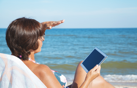 Woman with tablet pc at the beach. Elderly woman in a swimsuit sunbathing and holding a tablet on summer sea beachの写真素材