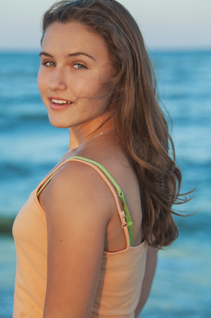 Young long brown-haired teen girl looking at camera and smiling on beach at summer eveningの写真素材