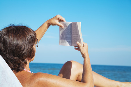 Elderly woman in a swimsuit sunbathing and reading a book on summer sea beach. beautiful woman reading a book on background of beach.の写真素材