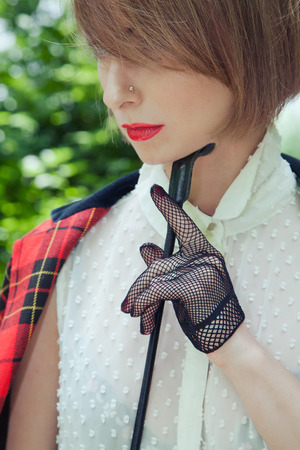Close-up portrait of beautiful young woman in horsewoman suit in green forest on sunny day. Girl holds riding stack in her hand and stroking it her chinの写真素材
