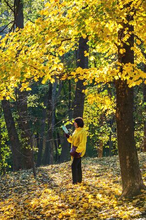 Adult woman in yellow jacket standing in fallen leaves of autumn forest and readingの写真素材