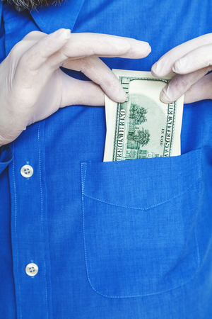 Hand in rubber glove hides in the breast pocket of a blue uniform shirt dollar bill. Close-upの写真素材