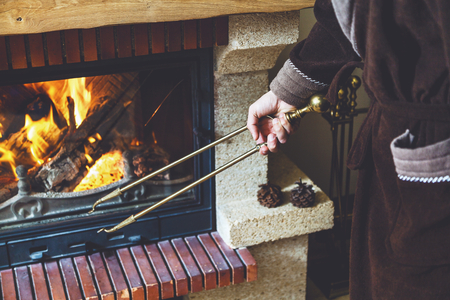 A man in a warm robe holding a large brass fireplace tongs. In a fireplace fire burns brightの写真素材