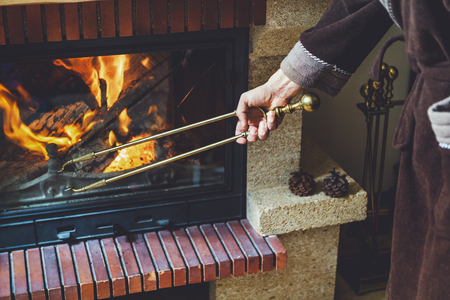 A man in a warm robe holding a large brass fireplace tongs. In a fireplace fire burns brightの写真素材