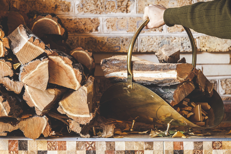 hand of man holding curved handle copper woodpile filled with firewood. Nearby on a shelf stacked logsの写真素材