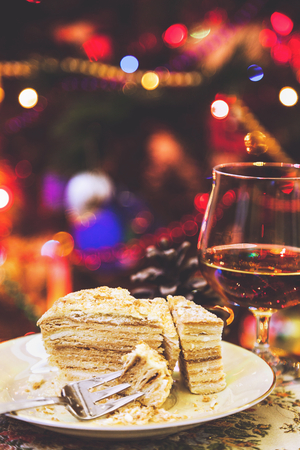 Holiday still life. Piece of cake and dessert fork rests on a plate close-up. In the background, an elegant glass of cognac and a bump. Behind blurred lights Christmas tree garlands
の写真素材