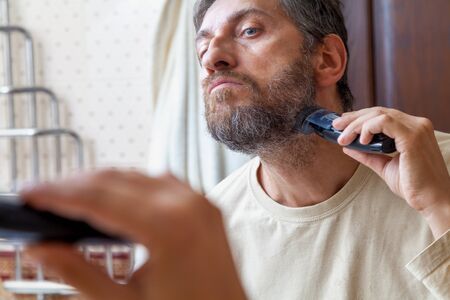 Grooming a beard trimmer. A nice gentleman in a beige T-shirt yourself shear his gray beard with the...