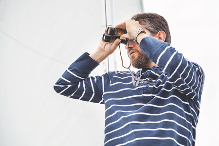 Cute man with a beard with a striped shirt and looking through binoculars sailing yacht closeup