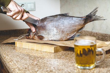 Cutting dried fish. Hands cuts a large dried-down bream with a large knife. Nearby is a mug of beerの写真素材