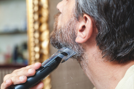 Grooming a beard trimmer. Man cuts his own gray beard using a typewriter closeupの写真素材