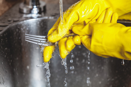 Washing dishes. Hands in yellow rubber gloves wash a large spoon under a stream of running water in a metal sink close-upの写真素材