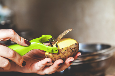 Kitchen work. The hands of a woman with a manicure brush potatoes with a plastic tool with a special blade close-up. Side viewの写真素材