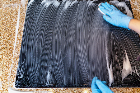 Hands in blue rubber gloves wash the glass surface of the electric oven with a soft sponge. Is...