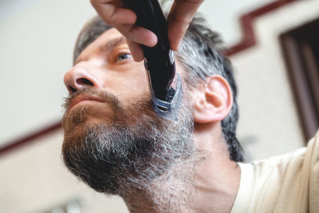 Grooming a beard trimmer. A nice gentleman in a beige T-shirt yourself shear his gray beard using a typewriter view from belowの写真素材