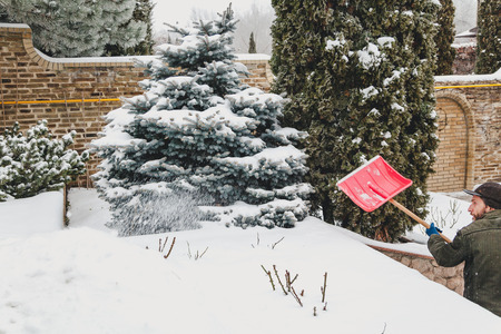 man with a beard brushing the snow red plastic shovel in the yard of a private house. On the terrace, grow treesの写真素材