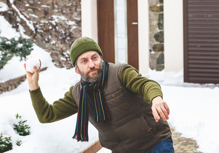 Snow games on the air. A man with a beard in a warm waistcoat and hat throws snowballs in the courtyard of a private house in a frosty day
の写真素材