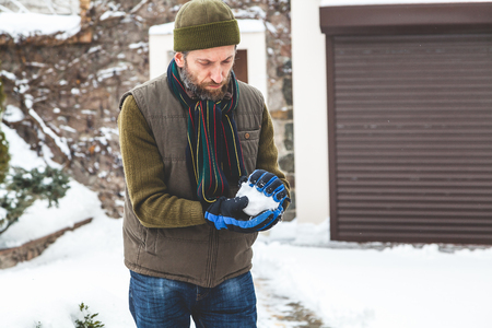 Winter fun. A man with a beard in a warm waistcoat and hat forms snowballs in the courtyard of a private house in a frosty day
の写真素材