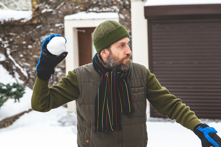 Snow games on the air. A man with a beard in a warm waistcoat and hat throws snowballs in the courtyard of a private house in a frosty dayの写真素材
