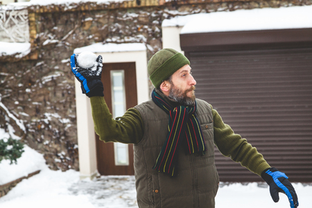 Snow games on the air. A man with a beard in a warm waistcoat and hat throws snowballs in the courtyard of a private house in a frosty day
の写真素材