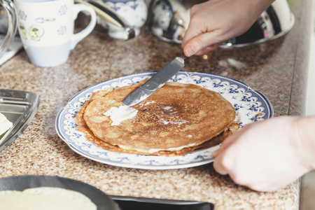 Hands grease appetizing fried pancake with butter with a table knife. A pile of pancakes lies on a blue plate
の写真素材