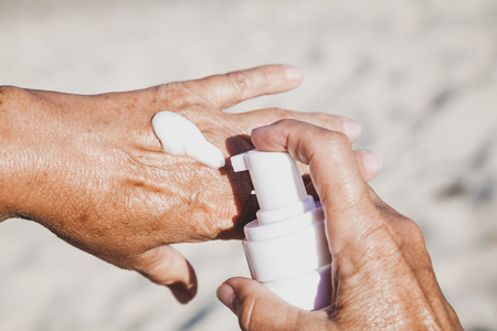 Elderly woman smears her hands with sunscreen from a container on the summer sea beachの写真素材