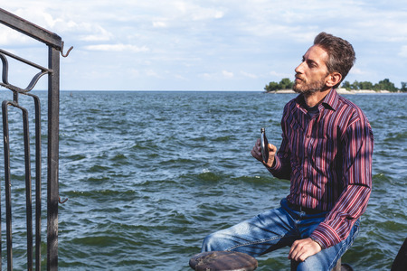 man with a beard sitting on the mooring bollard on a concrete pier and holding a flat flask on the bank of a wide river
の写真素材