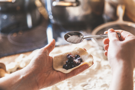 woman hand pours sugar into a raw dumpling with a stuffing of black round berries. On the table lie raw dumplings, sieve and rolling pin
の写真素材
