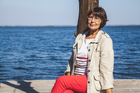Woman on the river bank. Cute elderly woman sitting on a blue water river background in a sunny dayの写真素材