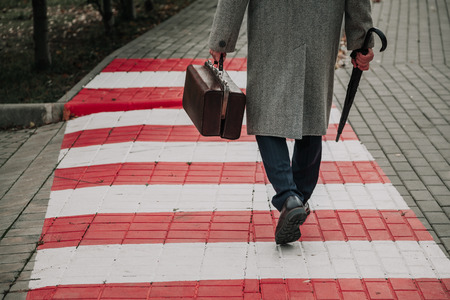 man in stylish shoes and a white coat walks along the red and white stripes of the pedestrian...