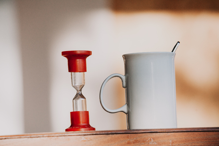 Time to drink tea.Strict high white cup with comfortable handle and hourglass on blurred background