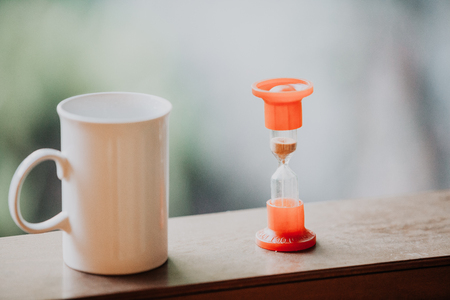 Time to drink tea.Strict high white cup with comfortable handle and hourglass on blurred backgroundの写真素材