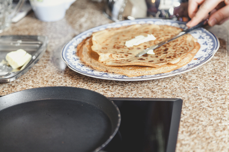 Hands grease appetizing fried pancake with butter with a table knife. A pile of pancakes lies on a blue plateの写真素材