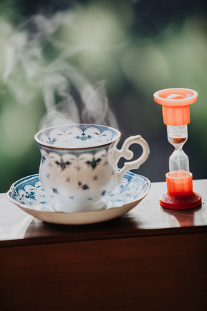 Time to drink tea. A neat cup for tea with a blue pattern and a saucer stand on wooden rails. Near the hourglassの写真素材