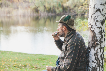 Rest on the shore. A man in the helmet of a deer hunter leans on the trunk of a birch and drinks...