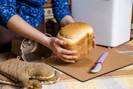 womans hands hold fresh bread from the bread baking mold of the electric bread maker. There is a big...