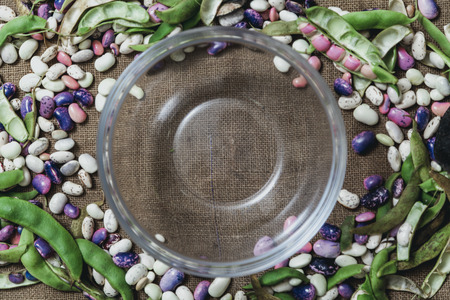 Pods and seeds of beans of different colors lie on the sacking along the edges. In the middle there...