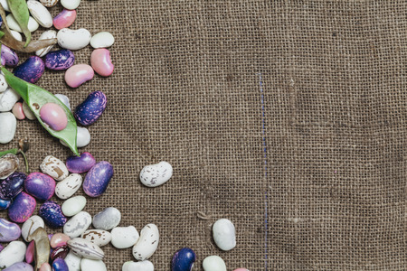 Pods and seeds of beans of different colors lie on burlap. View from above