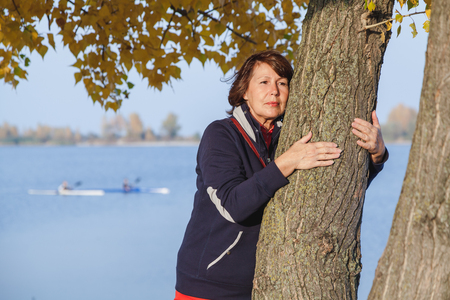 Cute elderly woman hugging a tree trunk on the shore of a pond on an autumnal sunny day closeupの写真素材
