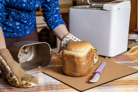 Womans hands in oven mitts pull fresh bread from the mold for baking bread electric bread maker.の写真素材