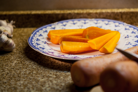 Pieces of chopped carrots lie on a plate. Next on the marble table is garlic and a whole carrotの写真素材
