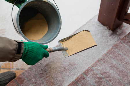 Injury prevention. Hand in green glove sprinkles sand on a winter path with a small shovel and bucketの写真素材