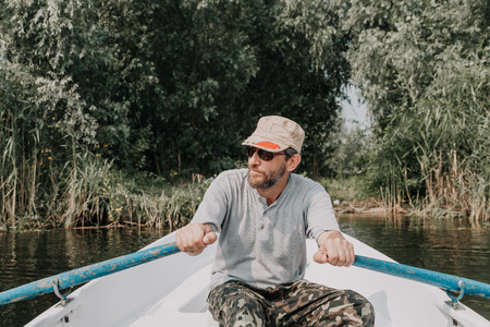 man with a beard in glasses and camouflaged trousers controls a rowing boat in the middle of the river on a clear dayの写真素材