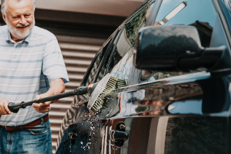 gracious man with a gray beard washes a black car with a special brush on a long handleの写真素材