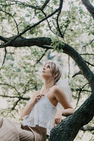 Portrait in nature. Slim stylish girl in a white top sitting on a tree branch on a summer dayの写真素材