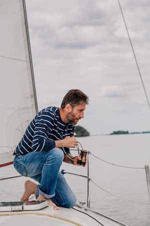 man with a beard in a striped sweater and jeans sits on the bow of a sailing yacht and looks overboard on a cloudy day.の写真素材