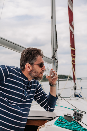 Businessman with a beard in dark glasses and a striped sweater stands on a white yacht and drinks whiskey from a glassの写真素材
