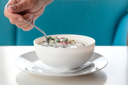 Appetizing summer soup in a round white plate close-up. Hand holds a tablespoon steel spoon