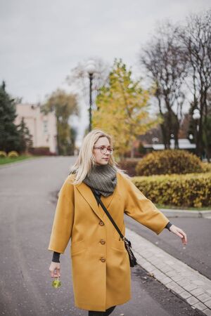 beautiful blonde in glasses and walking in an autumn park on her bright yellow fashion coatの写真素材